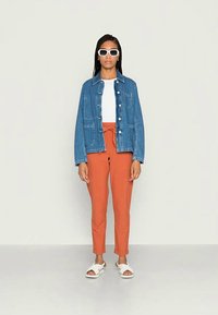 Casaco de ganga com bolsos frontais, t-shirt branca, calças laranja com cordão e sandálias brancas. O modelo usa óculos de sol redondos brancos.