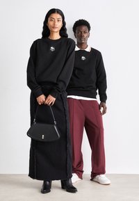 Sweat-shirt noir avec logo blanc, jupe frangée noire, sac à main noir, pantalons bordeaux et baskets blanches. Design minimaliste et tissus texturés.