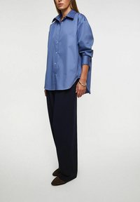 Blå satinskjorta med klassisk krage, långärmad och knäppning framtill, kombinerad med löst sittande mörka byxor och platta skor.