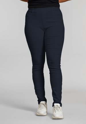 Osoba w granatowych, dopasowanych spodniach i białych trampkach, stojąca na tle jednolitego jasnoszarego tła.