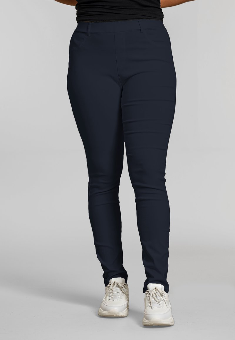 Isik, kes kannab kitsaid tumesiniseid pükse ja valgeid tenniseid, seisab heleda halli ühevärvilise tausta ees.