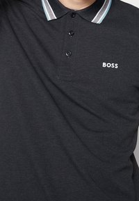 Černé polo tričko s límečkem a pruhovanými detaily, tříknoflíkovou léga a vyšitým logem 'BOSS' na hrudi.