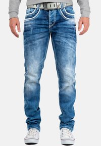 Jeans in denim blu chiaro con un look scolorito, caratterizzati da cuciture bianche e cinque tasche, abbinati a un maglione grigio e scarpe bianche.