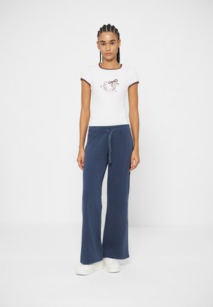 Jeune femme aux cheveux tressés portant un t-shirt blanc avec un motif fer à cheval, un pantalon bleu marine à cordon de serrage et des baskets blanches, debout devant un fond uni.
