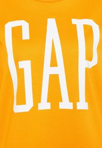 Gul bomulls-t-shirt med stor vit "GAP"-logotyp på framsidan. Mjuk textur med rund halsringning.