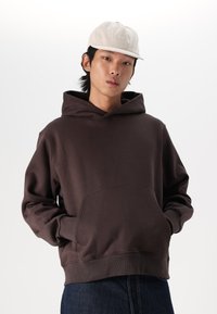 Dark brown hoodie with a front pocket, featuring a spacious hood. The model also wears a light beige cap with the brand logo.