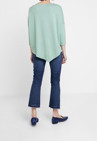 Maglia oversize verde chiaro con maniche a tre quarti, abbinata a jeans a zampa d'elefante blu scuro e ballerine marine. Tessuto texturizzato e orlo irregolare.