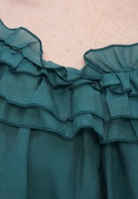 Τοπ από τιρκουάζ μουσελίνα με στρωματοποιημένες βολάν στο λαιμό, με λεπτές ραφές κατά μήκος των άκρων και απαλή, διαφανή υφή.