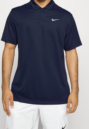 Homme portant un polo Nike bleu marine à manches courtes et un short blanc Nike, debout devant un fond clair uni.
