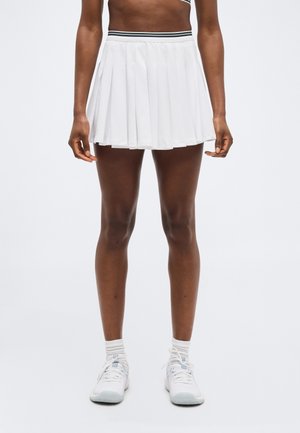 Personne portant une jupe de tennis plissée blanche avec une ceinture rayée, des chaussettes blanches et des chaussures de sport blanches, debout devant un fond uni.