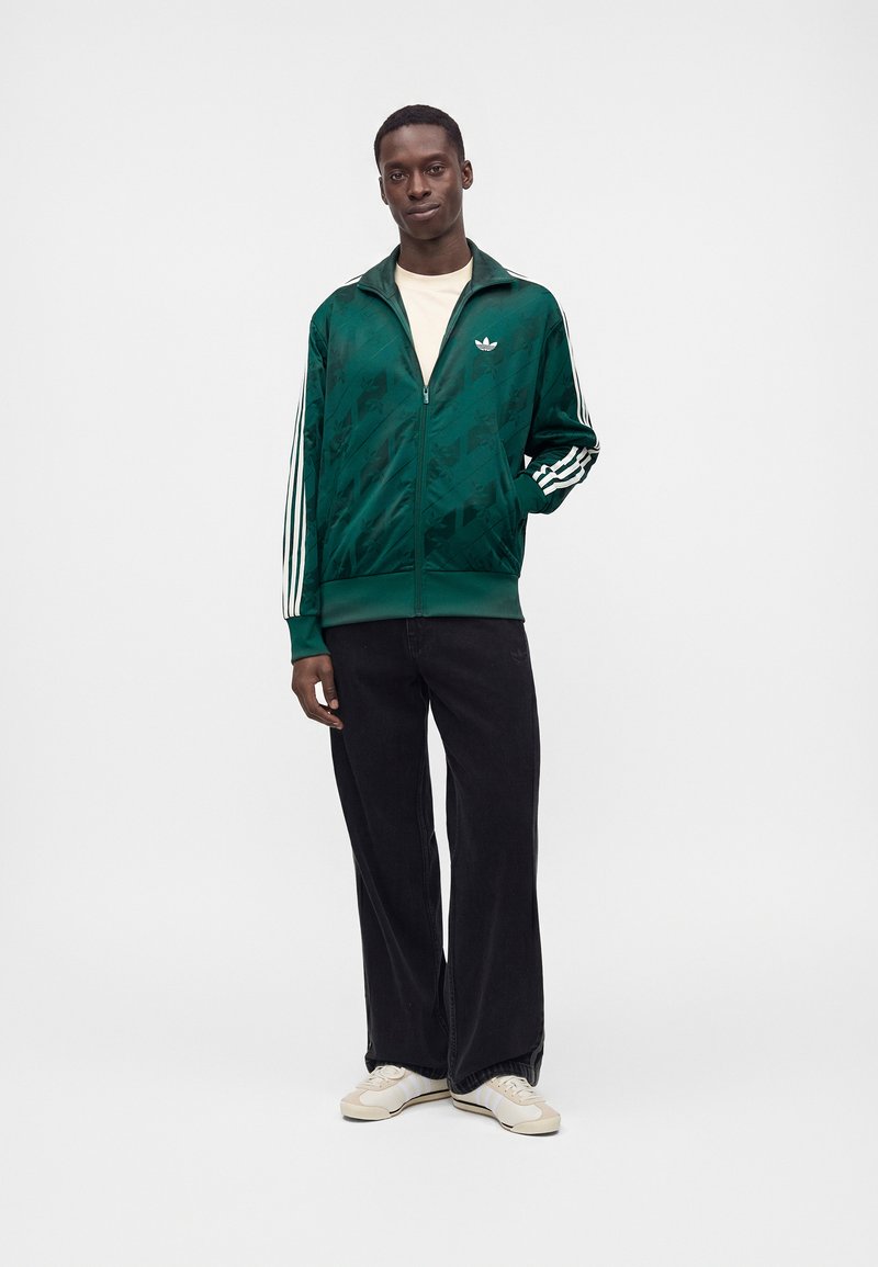 Hombre que lleva una chaqueta Adidas verde oscuro con cremallera y rayas blancas en las mangas, pantalones negros de pierna ancha y zapatillas blancas, de pie frente a un fondo liso.