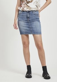 Jupe en denim mini en délavage bleu moyen, avec cinq poches, un design ajusté et des coutures contrastées. Associée à des bottines noires.