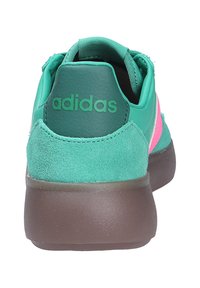 Zelena tenisica s teksturama od antilopa i kože, s ružičastom prugom, zaobljenom stražnjom stranom i gumenim potplatom. Ime brenda "adidas" ispisano u zelenoj boji.