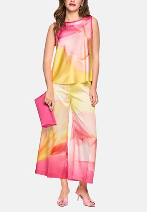 Frau trägt ärmelloses Oberteil und weite Hosen mit abstraktem Blumenmuster in Rosa, Gelb und Orange, hält eine rosa Clutch und passende Schuhe.