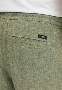 Zelene herringbone vzorčne hlače s hrbtnim žepom in črno oznako, na kateri piše "POSVEČENO DENIMSKI POPOLNOSTI." Ohlapna zasnova.
