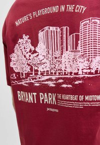 Mørkerød bomulds-T-shirt med et pink grafisk motiv af Bryant Park med træer og skyskrabere, fed tekst og et tekstureret design.