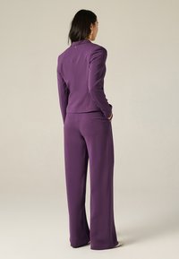 Completo viola composto da un blazer aderente e pantaloni a gamba larga realizzati in tessuto morbido, caratterizzati da una sottile cucitura decorativa e tasche laterali.