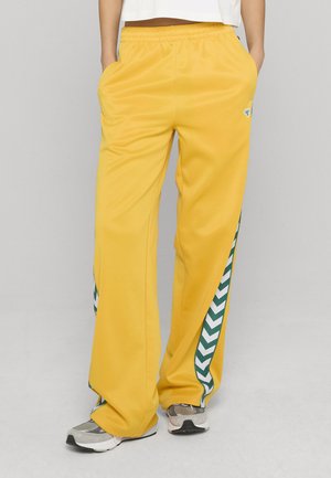 Žluté polyesterové tepláky s proužky na bocích v zelenobílém cikcak vzoru. Mají elastický pas a přední kapsy.