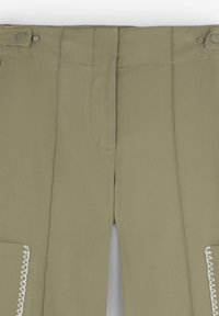 Hellekhakifarbene Hose mit hoher Taille, ausgestattet mit einem Reißverschluss vorne, zwei Taschen und dekorativen weißen Nähten entlang der Taschenkanten.