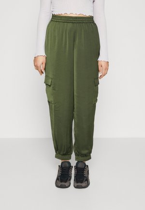 Persoon draagt olijfgroene cargobroek met elastische boorden en taille, een witte cropped top met lange mouwen, en zwarte en grijze sneakers.