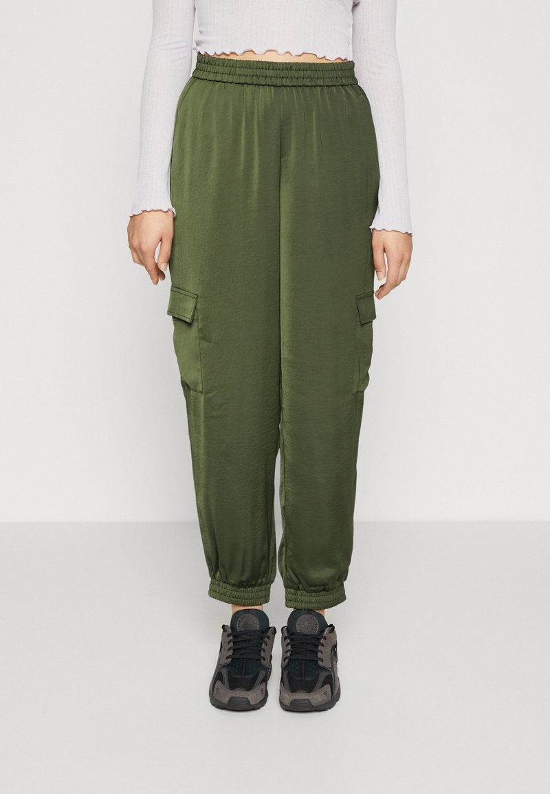 Personne portant un pantalon cargo vert olive avec des poignets et une taille élastiques, un crop top blanc à manches longues, et des baskets noires et grises.