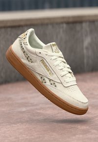 Tenisky Reebok s krémovým semišovým zvrškom, akcentmi s leopardím vzorom, bielymi šnúrkami a hnedou gumovou podrážkou. Prítomné zlaté logá.