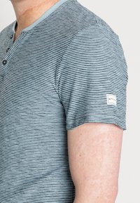 Pruhovaná kratší halenka s krátkým rukávem v tlumené tyrkysové a černé barvě. Obsahuje zapínání na knoflíky v přední části a boží štítek; měkká, lehká tkanina.