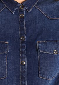 Dunkelblaue Jeanshemd mit durchgehender Knopfleiste, Kragen und zwei Brusttaschen mit Messingakzenten und kontrastierenden Stichen.
