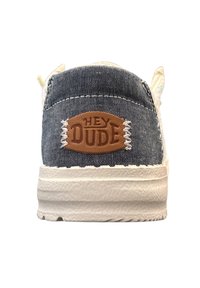 Canvas sneakers met een blauwe bovenkant, witte zool en een bruine lederen tag met de tekst "HEY DUDE" op de hiel. Textuurdetail aanwezig.