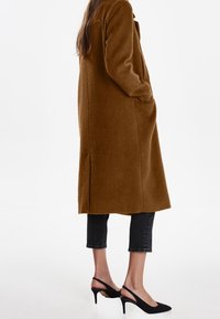 Brun uldlang frakke med en blød tekstur, forlommer, parret med sorte hælede slingback-sko og mørke cropped jeans.