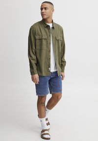 Olijfgroen overhemd met knoopsluiting en twee borstzakken, gedragen over een wit T-shirt. Gecombineerd met blauwe denim shorts en bruine sandalen.