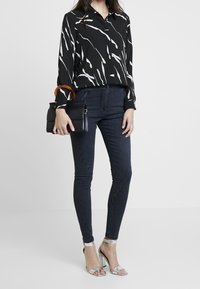 Schwarzes Button-Up-Hemd mit weißen abstrakten Linien, kombiniert mit dunklen Skinny-Jeans und metallischen High-Heels. Schwarze Handtasche mit transparentem Griff.
