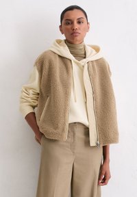 Beige Shearling-Weste mit einem Reißverschluss vorne, getragen über einem cremefarbenen Hoodie und einem beigen Rollkragenpullover, kombiniert mit beigen weit geschnittenen Hosen.