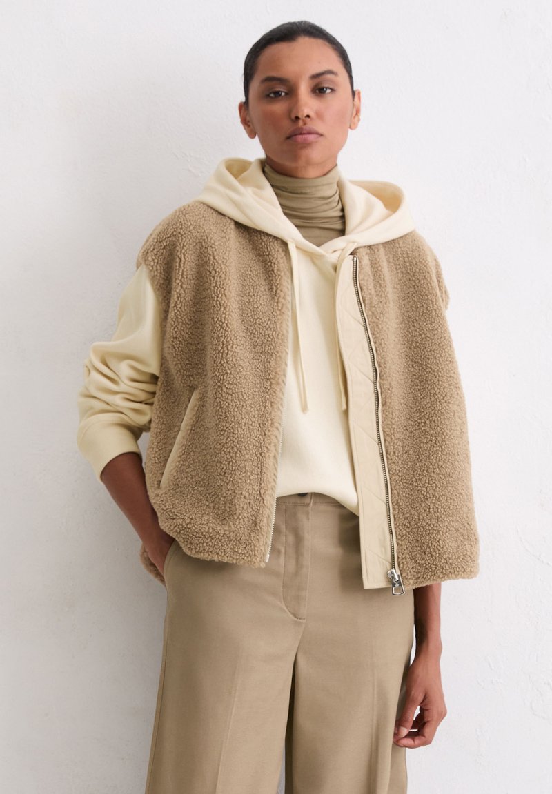 Beige Shearling-Weste mit einem Reißverschluss vorne, getragen über einem cremefarbenen Hoodie und einem beigen Rollkragenpullover, kombiniert mit beigen weit geschnittenen Hosen.
