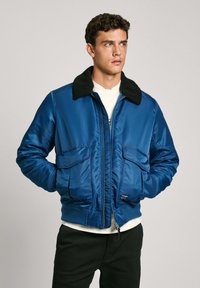 Chaqueta bomber azul con un cuello negro texturizado, cierre frontal con cremallera y dos bolsillos frontales. Fabricada en un material suave y brillante con puños acanalados.