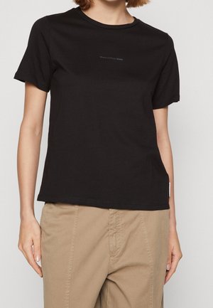 Kvinde iført en enkel sort t-shirt med korte ærmer og lille "Marc O'Polo DENIM" tekst på brystet samt beige bukser.