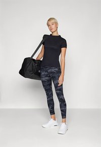 Camiseta deportiva negra, leggings con estampado de camuflaje en tonos grises y azul oscuro, zapatillas blancas y una bolsa de deporte negra.
