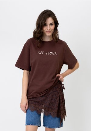 Ung kvinde med skulderlangt brunt hår iført en brun t-shirt med teksten "OH APRIL" og en blondernederdel over cowboyshorts.