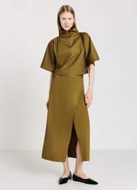 Top corto verde oliva con colletto alto e tasca frontale, abbinato a una gonna avvolgente coordinata con uno spacco laterale. Completa il look un paio di ballerine nere.