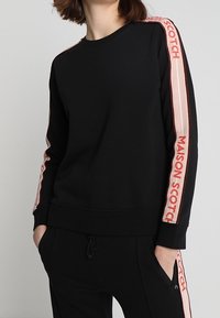 Czarna bluza z miękkiego materiału, z długimi rękawami w różowe i czerwone paski oraz napisem "MAISON SCOTCH" po bokach.