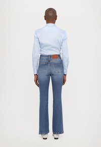 Svijetloplena košulja s ovratnikom i zatvaranjem na gumb, uparena s trapericama visokog struka u srednje plavoj boji. Smeđi kožni detalj na trapericama.