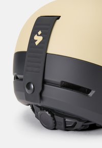 Kask rowerowy z beżową zewnętrzną powłoką i czarną podstawą. Obejmuje teksturowany pokrętło do regulacji oraz bezpieczny pasek z detalem logo.