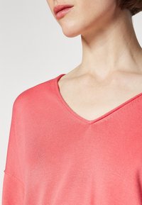 Femme portant un pull en maille corail rose à col en V, recadré pour montrer le cou et les épaules sur un fond clair uni.