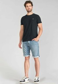 T-shirt en coton noir, short en denim bleu clair avec des ourlets retroussés, et baskets noires et blanches. Le modèle pose avec une main dans la poche.