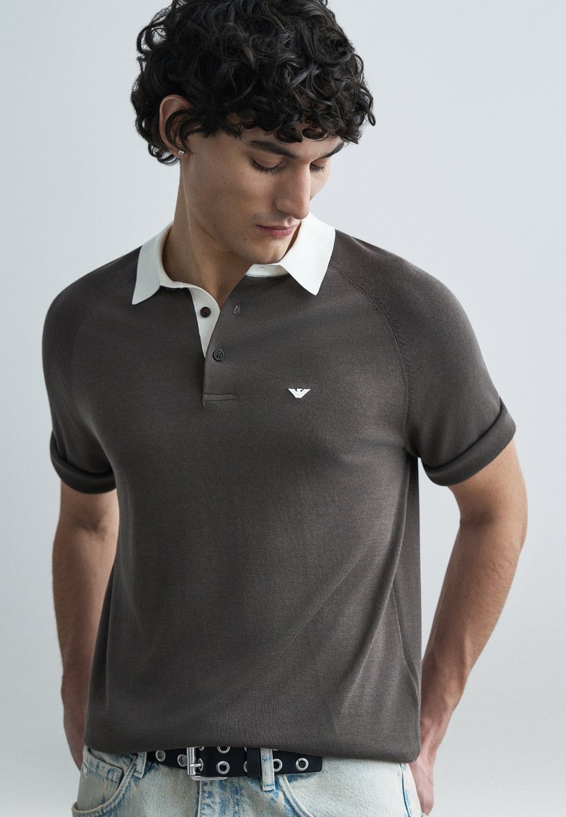 Polo marron avec un col blanc, des manches courtes et un petit logo sur la poitrine. Le tissu a une texture lisse et une coupe ajustée.