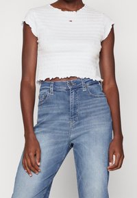Bílý smockovaný crop top s kulatým výstřihem a volánkovými okraji, kombinovaný se světle modrými džíny s vysokým pasem, které mají pět kapes.