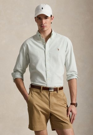 Fehér baseball sapkát viselő férfi, világos csíkos ing, felhajtott ujjú, barna öv, őzszínű rövidnadrág és karóra; semleges háttér előtt áll.