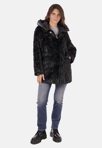 Cappotto in pelo sintetico nero con cappuccio removibile, dotato di zip e chiusura a pressione, abbinato a jeans blu e stivali neri.