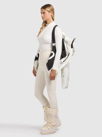 Haut blanc à manches longues, leggings crème et bottes isolantes blanches. Elle tient une veste matelassée noire et blanche, avec fermetures éclair et sangles.