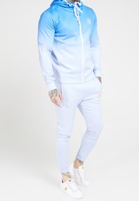 Tuta blu chiaro con un design a gradiente, zip frontale e cappuccio. I pantaloni slim fit hanno tasche laterali e un dettaglio con logo. Le sneakers bianche completano il look.